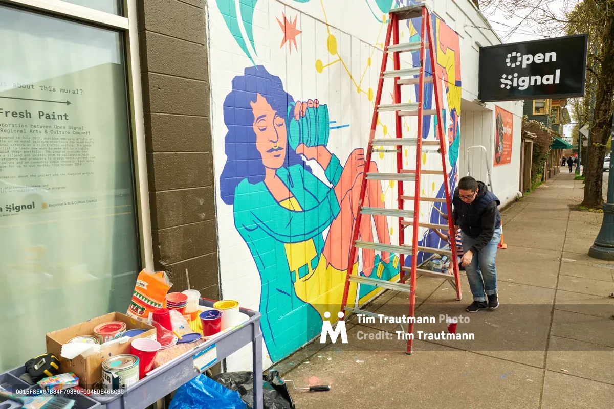 Illustrator Anke Gladnick works methodically on her vibrant mural "Let's Talk" outside Open Signal on Northeast Martin Luther King Jr. Boulevard in Portland's historic Albina neighborhood. The artist crouches beside a red ladder, applying paint to the lower portion of her surreal composition featuring a figure with flowing blue hair holding a large teal camera or recording device. Paint containers and brushes are scattered across a makeshift table, while the afternoon light illuminates both the emerging artwork and the urban streetscape of this culturally significant corridor.