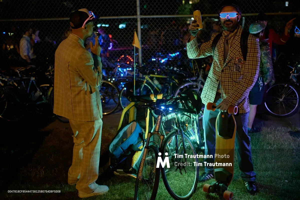 Two costumed cyclists pause beneath the glowing chain-link boundary of Colonel Summers Community Garden during Portland's Drop Out Prom Ride, part of the city's celebrated Pedalpalooza festival. The man on the right sports a checkered blazer and illuminated shutter shades while holding what appears to be a beer, embodying the event's playful anti-establishment spirit. Neon lighting from decorated bicycles casts an electric blue-green glow across the nighttime scene, creating an atmosphere where formal wear meets cycling counterculture.
