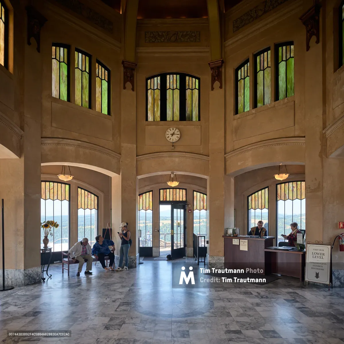 The grand interior of Vista House at Crown Point reveals its majestic Art Nouveau architecture, with soaring limestone walls and distinctive stained glass windows filtering golden afternoon light. Visitors gather beneath the ornate two-story rotunda, some seated on benches while others explore the information desk, creating an intimate human scale against the monument's imposing classical proportions. The polished marble floors reflect the warm illumination streaming through the geometric leaded glass panels, while a vintage clock marks time in this beloved Oregon landmark overlooking the Columbia River Gorge.