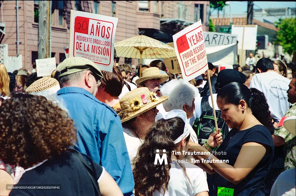 A dense crowd of Mission District residents gathers in passionate demonstration against displacement, their weathered faces and raised protest signs creating a mosaic of community resistance. Bilingual placards declaring "30 AÑOS RESIDENTE ORGULLOSO DE LA MISIÓN" and "4 YEARS PROUD RESIDENT OF THE MISSION" punctuate the sea of sun hats and determined expressions. The warm afternoon light filters through the urban streetscape of Bryant Street, illuminating this grassroots mobilization where longtime neighbors stand shoulder to shoulder, defending their cultural heritage against the encroaching tide of tech gentrification.
