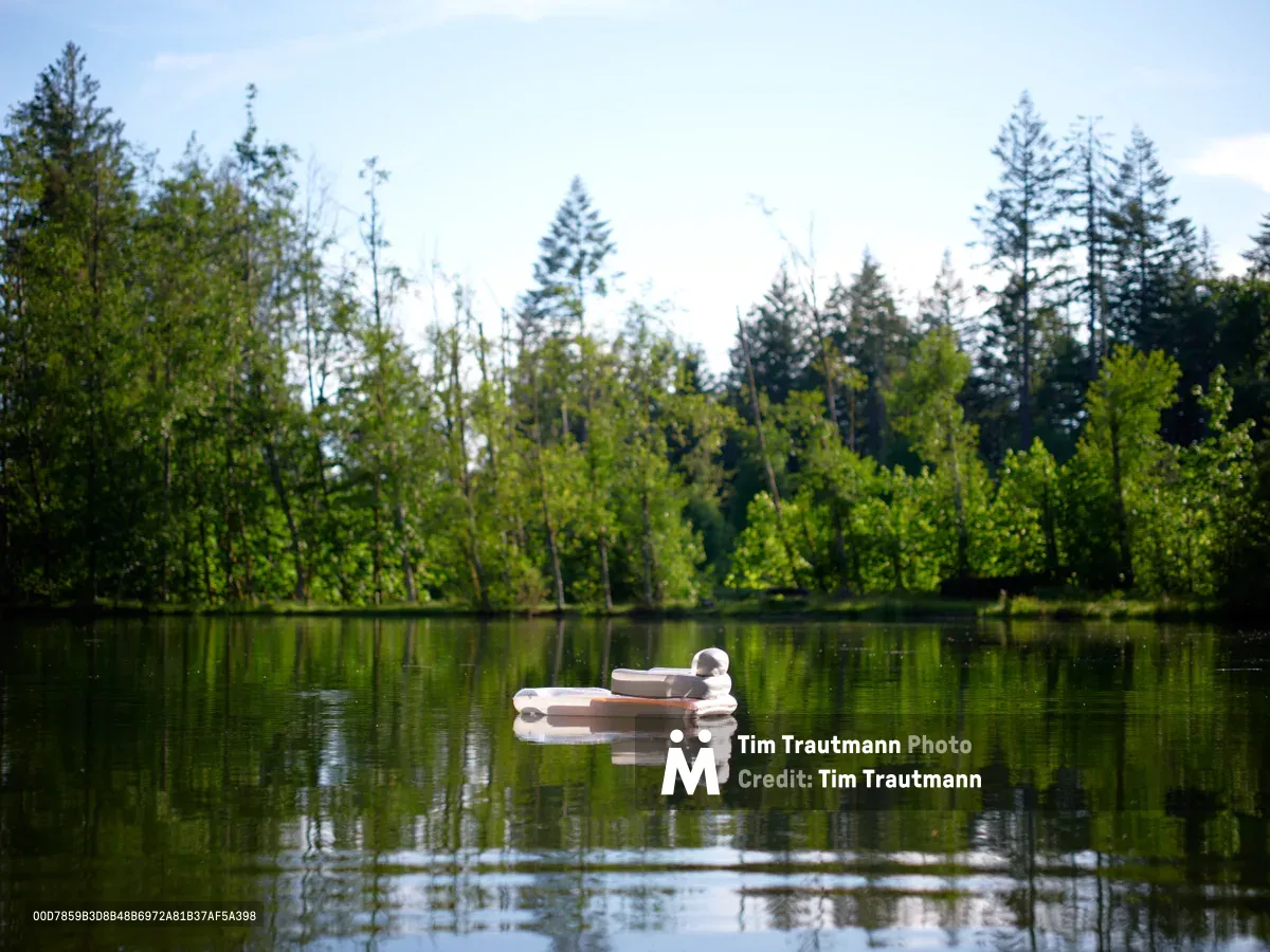 An inflatable raft drifts serenely across the glass-like surface of a tranquil lake, its form creating a perfect reflection in the still water. The scene is framed by a verdant shoreline where fresh spring foliage in brilliant greens creates a lush canopy, while tall conifers punctuate the background against a soft, overcast sky. The composition captures a moment of pure quietude, with the raft positioned as a gentle focal point within this pristine natural sanctuary.