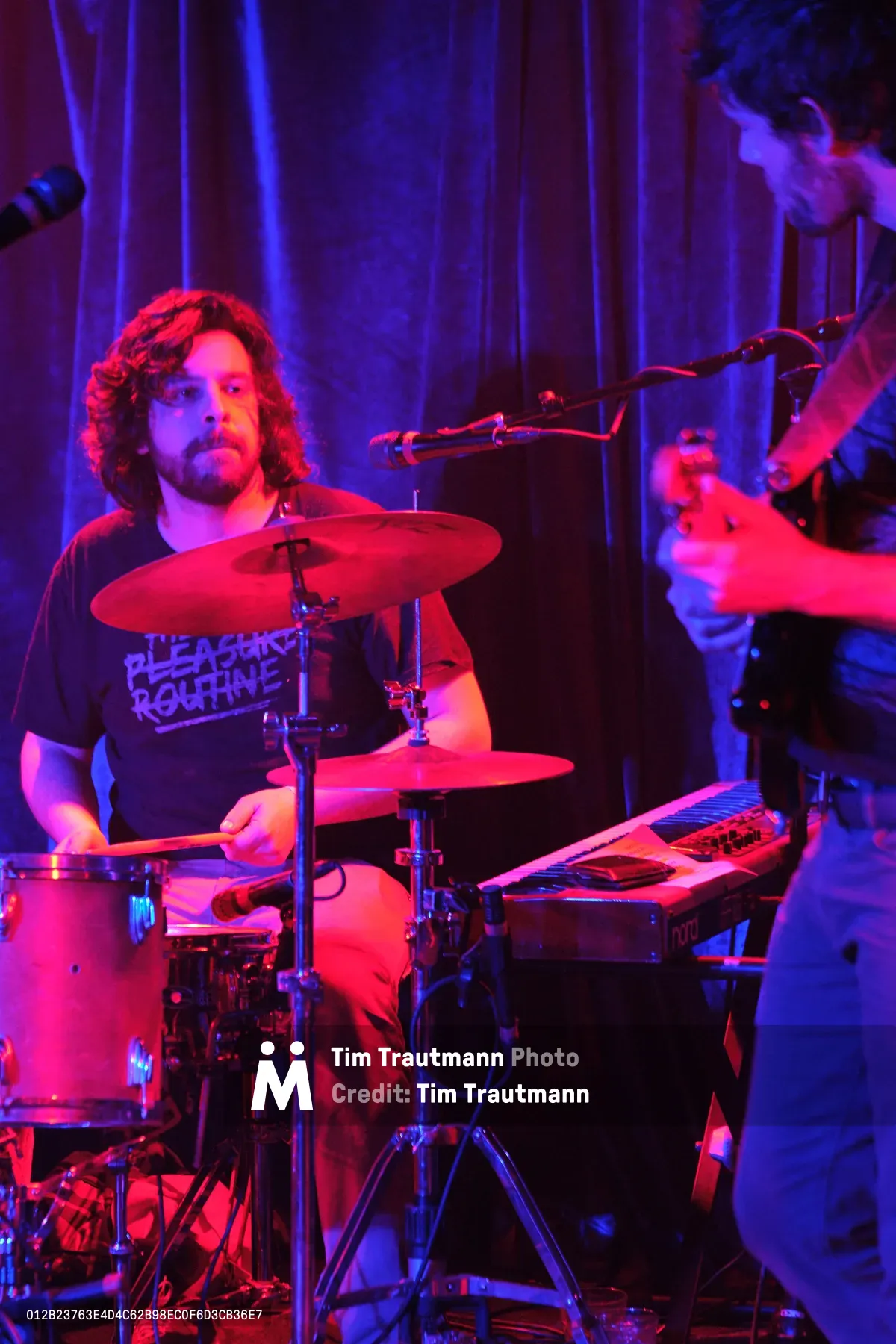 A curly-haired drummer from Sun Blood Stories commands the kit at Portland's legendary Dante's venue, his form bathed in intense red and purple stage lighting. The atmospheric illumination transforms the intimate club setting into a moody sonic sanctuary, with vertical light beams cutting through the haze behind him. His bandmate's silhouette lurks in the background shadows, creating depth in this electrifying live performance moment.
