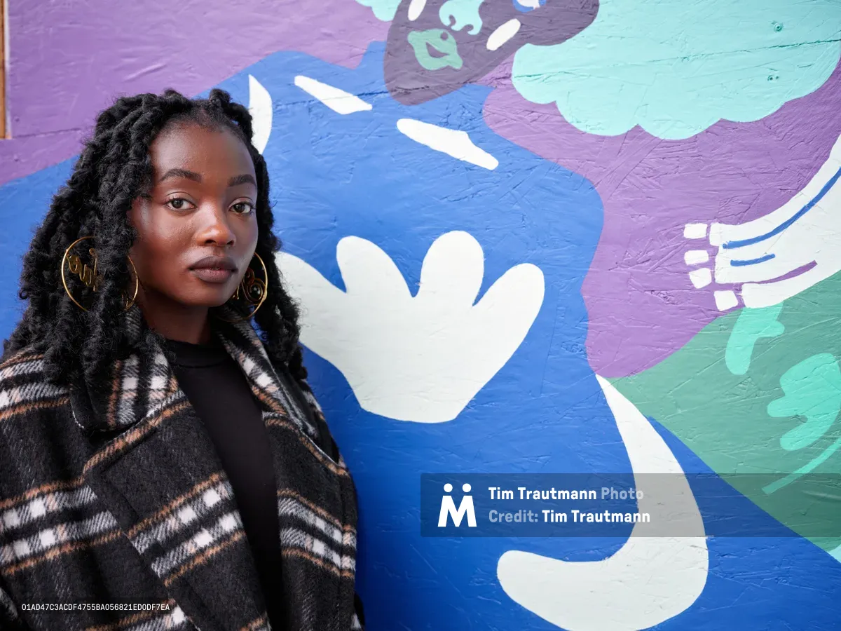 Artist Naomi Likayi poses thoughtfully before her vibrant mural adorning the boarded World Trade Center in Portland, Oregon. Her protective locs frame her face as golden hoop earrings catch the natural light, while she wears a striking plaid wool coat over a black top. The abstract mural behind her explodes in oceanic blues, soft purples, and mint greens, featuring bold white hand silhouettes that seem to reach across the painted surface with gestural energy.