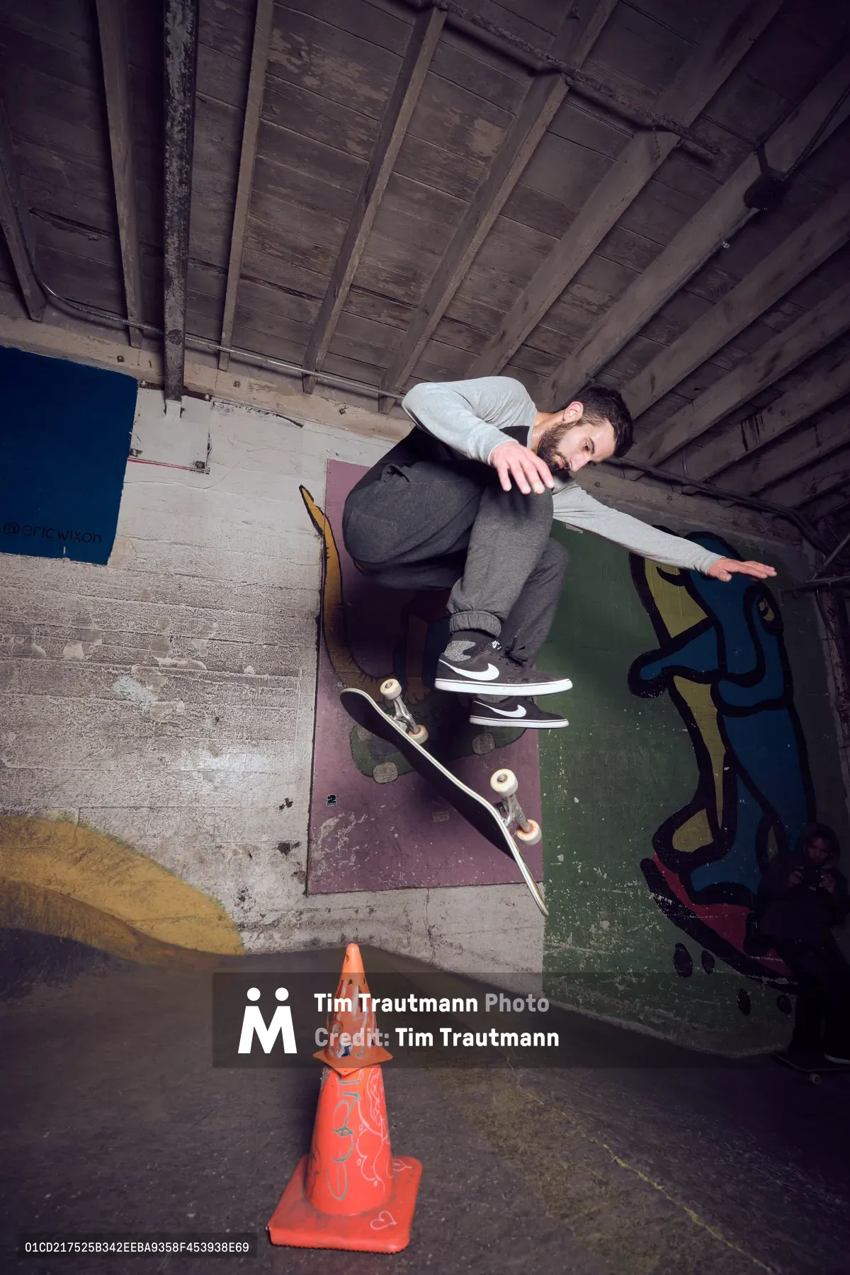 A skateboarder poses confidently in the raw industrial interior of Commonwealth Skateboarding in Portland's Buckman neighborhood. The young man sits casually against weathered concrete steps beneath exposed wooden ceiling beams, his skateboard resting nearby alongside a bright orange traffic cone. Moody lighting illuminates the gritty urban architecture, capturing the authentic underground skateboarding culture that defines Portland's Southeast 20th Avenue scene.