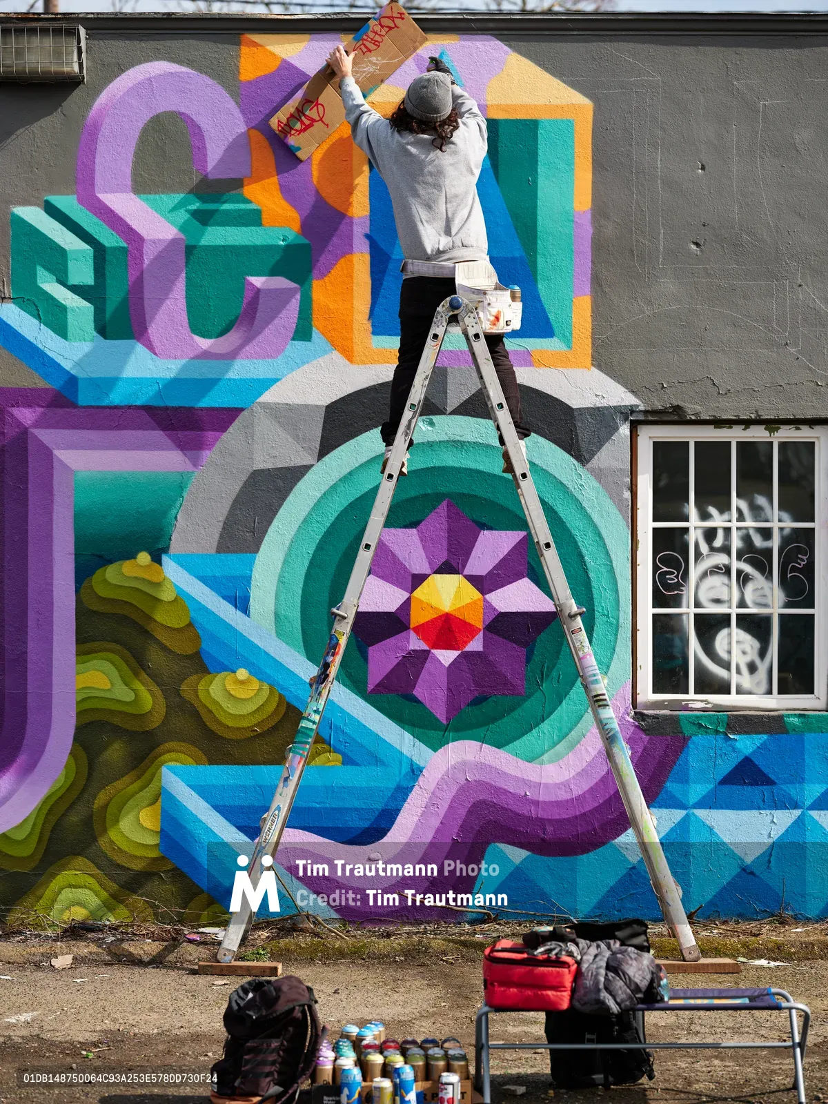 A muralist in gray hoodie and beanie balances precariously on a stepladder, adding finishing touches to a vibrant geometric wall painting in Northeast Portland, Oregon. The kaleidoscopic mural features bold dimensional typography and mandala-like patterns in electric purples, teals, oranges, and blues against weathered gray concrete. Spray paint cans and art supplies scattered below reveal the creative process, while diffused daylight illuminates the transformation of urban space into colorful public art.