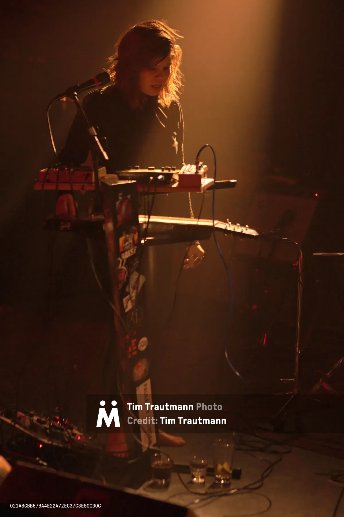 A female musician from experimental band Sun Blood Stories performs on keyboard at Dante's venue in downtown Portland, her silhouette dramatically lit by warm amber stage lighting that creates a halo effect around her tousled hair. She stands focused behind a multi-tiered keyboard setup, surrounded by the intimate atmosphere of the underground music scene. The moody lighting and shadowy backdrop capture the raw energy of this Boise-founded band's mesmerizing live performance.