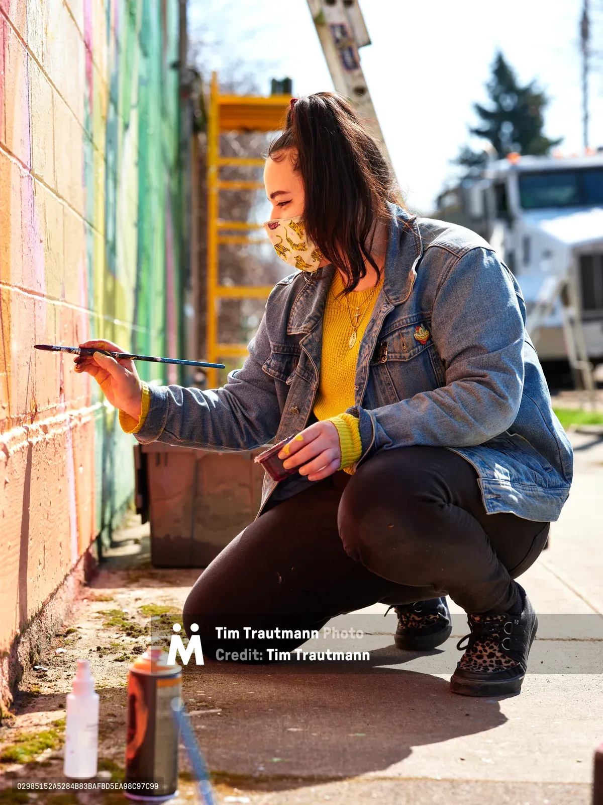 Multidisciplinary artist Vilatose crouches beside a weathered brick wall on Northeast Alberta Street, delicately applying paint to her emerging mural titled Jardín Cubana, which depicts three female figures. Golden afternoon light illuminates her yellow sweater and denim jacket as she works with focused concentration, her patterned face mask a contemporary reminder of the times. Spray paint cans rest on the concrete sidewalk beside her, while the colorful urban landscape of Portland's Alberta Arts District provides a vibrant backdrop to this moment of creative expression.