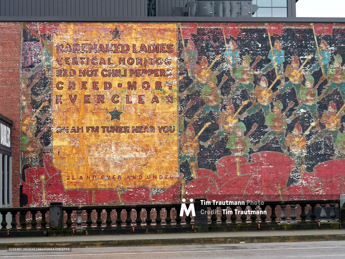 A weathered vintage billboard mural spans across a brick wall near Portland's Burnside Bridge, advertising alternative rock station KNRK 94.7 FM. The deteriorating paint reveals layers of amber, crimson, and charcoal hues, with band names like Barenaked Ladies, Red Hot Chili Peppers, and Everclear scattered across the composition. Industrial architecture looms above while an ornate balustrade frames the bottom of this urban archaeological artifact from the late 1990s alternative rock era.