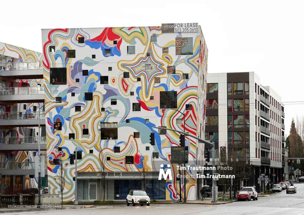 A striking apartment building in Portland's Buckman neighborhood showcases an extraordinary psychedelic mural that completely envelops its facade. The massive artwork features flowing ribbons of vibrant blues, reds, yellows, and earth tones that cascade across the white surface, punctuated by geometric black squares positioned over windows. The overcast Pacific Northwest sky creates a muted backdrop for this explosion of color, while modern residential buildings and bare winter trees frame the scene along the quiet urban street.