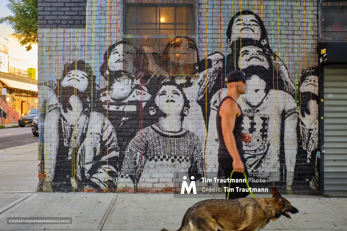 Golden hour light bathes a powerful mural on South 6th Street in Williamsburg, Brooklyn, featuring monochromatic portraits of cultural figures rendered in striking black and white against weathered brick. A man in athletic wear walks his German Shepherd past the towering artwork, creating a compelling juxtaposition between monumental street art and intimate neighborhood moments. The warm evening glow illuminates both the vibrant paint drips cascading down the wall and the peaceful domestic scene unfolding below.