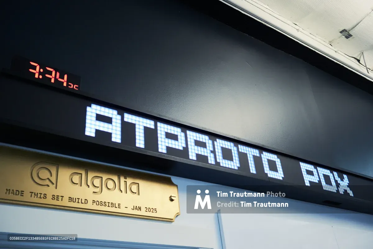 A pixelated LED display reading "ATPROTO POX" glows in cool blue light against dark industrial walls in Portland's CodeTV studio. The digital signage creates a stark technological contrast with the brass nameplate below bearing "algolia" and construction details from January 2025. Red time digits pierce the shadows above, while fluorescent ceiling lights cast harsh institutional brightness across this convergence of old and new tech aesthetics.