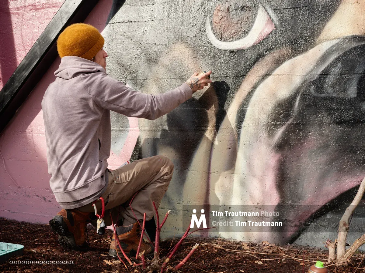 Artist Wetiko crouches in focused concentration, adding delicate details to a vibrant mural on a concrete wall in Portland's Hosford-Abernethy neighborhood. Wearing a mustard beanie and gray hoodie, the muralist works with precision against a backdrop of swirling pink, gray, and cream spray-painted forms. The intimate scene captures the methodical process of urban art creation, with the artist's shadow dancing across the textured wall surface as natural light illuminates the creative workspace.