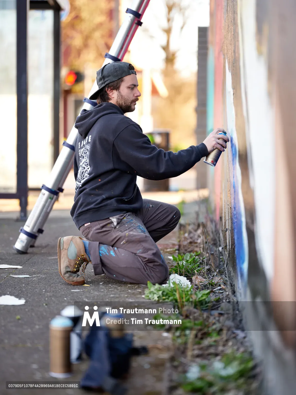 Travis Fields, known as campographic, kneels on the sidewalk applying spray paint to a mural of poet Amanda Gorman at the intersection of Northeast 15th Avenue and Killingsworth Street in Portland's Alberta neighborhood. The artist works in profile, his dark hoodie and backwards cap silhouetted against the warm afternoon light that bathes the urban streetscape. Paint cans scattered at his feet and a ladder positioned nearby speak to the methodical process of transforming this corner wall into public art.