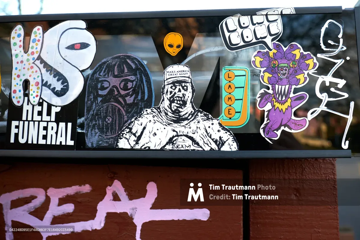 A collection of provocative stickers and street art adorns a black window frame on North Mississippi Avenue in Portland, creating a visual collage of contemporary political dissent. The eclectic mix includes cartoon characters, political imagery, and countercultural symbols plastered against the urban storefront, with additional pink graffiti marking the rust-colored wall below. The juxtaposition of playful imagery with darker political commentary captures the raw energy of Portland's street art scene in this gentrifying neighborhood.