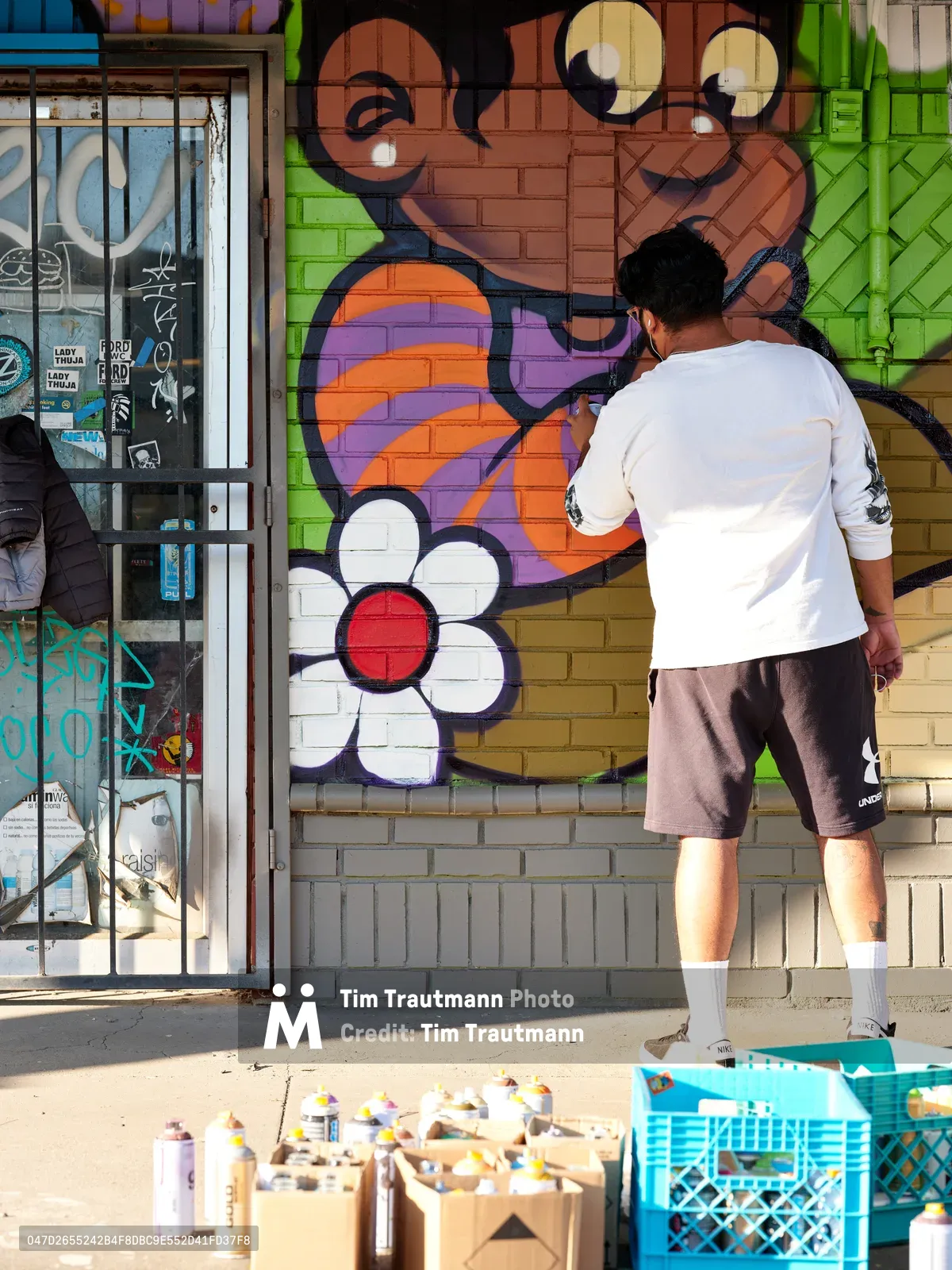 Muralist Heysus works intently on a colorful character mural featuring a cartoonish face with googly eyes and a simple white flower at the intersection of Killingsworth and NE 15th Avenue in Portland's Alberta district. Dressed casually in a white t-shirt and brown shorts, the artist applies spray paint to the brick wall while surrounded by crates of aerosol cans scattered across the concrete sidewalk. The afternoon light illuminates the vibrant oranges, purples, and greens of the emerging artwork against the urban storefront backdrop.