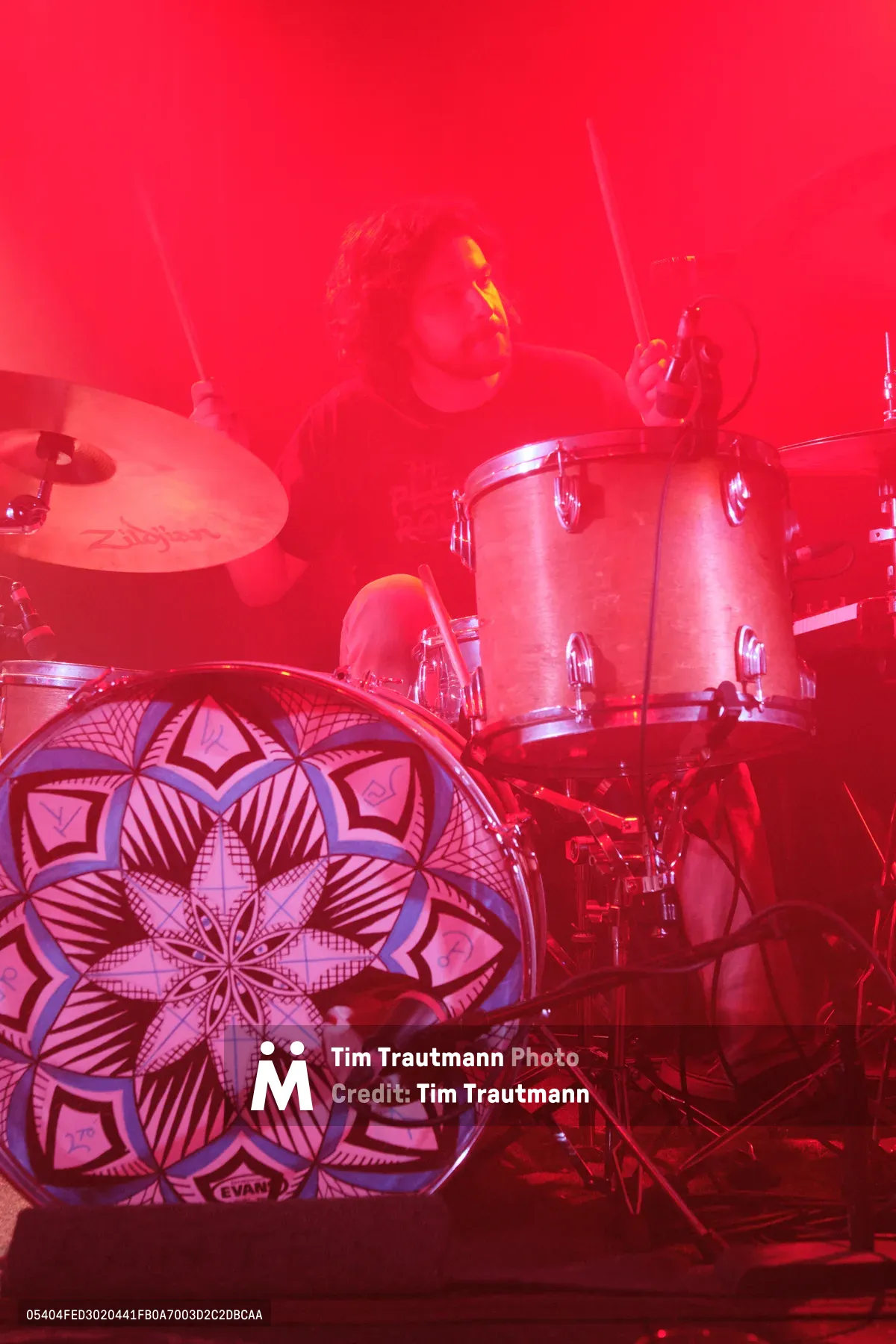 A drummer from Sun Blood Stories performs behind an ornate mandala-patterned kick drum, bathed in deep red stage lighting at Dante's in downtown Portland. The atmospheric lighting transforms the drum kit into a ceremonial altar, with intricate geometric patterns on the bass drum creating a mystical focal point. Cymbal stands rise like incense through the saturated crimson haze, while the performer remains partially obscured in the theatrical ambiance.