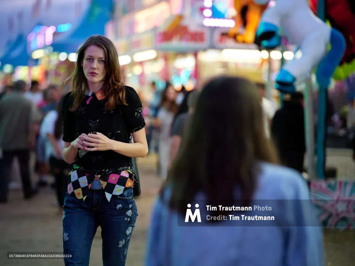 A young woman in a sequined black top and geometric belt stands amid the kaleidoscopic blur of carnival lights at the Clark County Fair in Vancouver, Washington. The shallow depth of field creates a dreamlike atmosphere as fairgoers drift past in soft focus, while vibrant neon signs and ride illumination paint the evening in electric blues, pinks, and amber. Her contemplative gaze and crossed arms suggest a moment of quiet reflection against the swirling energy of the midway.