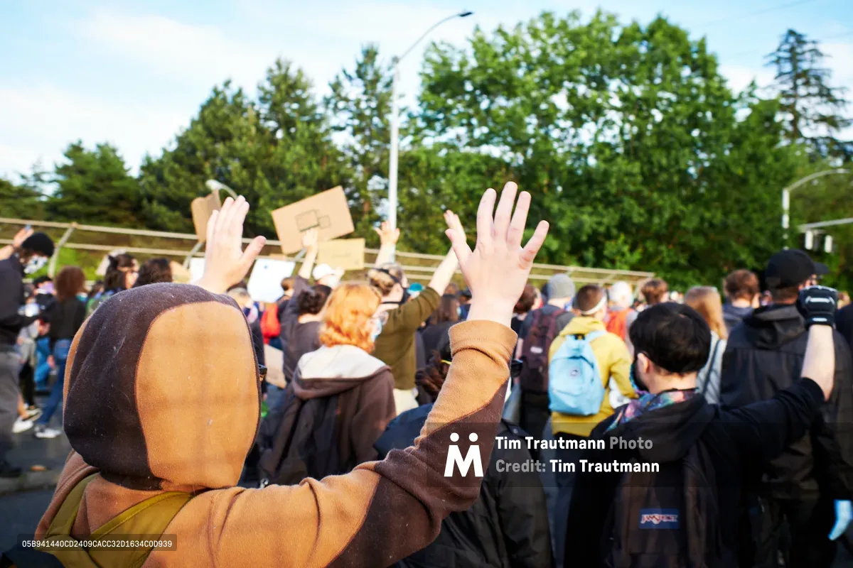 A demonstrator in a brown hoodie raises both hands skyward amid a crowded Black Lives Matter protest on Northeast 33rd Avenue in Portland's Sullivan's Gulch neighborhood. The scene captures the raw energy of collective action, with protesters holding cardboard signs against a backdrop of verdant summer trees and overcast skies. Golden hour light bathes the crowd, creating an atmosphere of both urgency and hope as voices unite in the aftermath of George Floyd's murder.