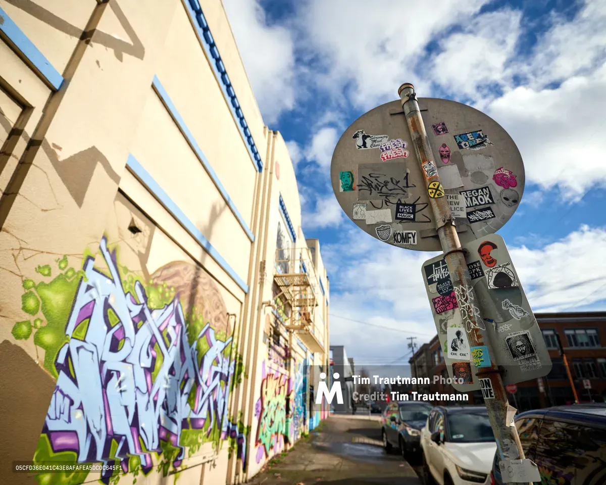 A vibrant graffiti mural dominates the cream-colored wall of a former Salvation Army warehouse in Portland's Central Eastside district, where purple and green wildstyle lettering creates a bold contrast against the weathered brick. A sticker-bombed street sign stands sentinel in the foreground, its circular surface layered with underground art collective logos and subcultural markers. The late afternoon light bathes the urban scene in warm tones while dramatic clouds sweep across the Pacific Northwest sky, creating an atmospheric backdrop to this commissioned street art installation.