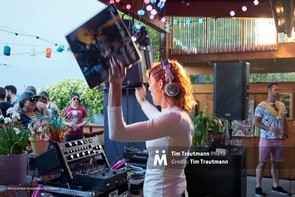 DJ Doctor Dakar, bathed in golden afternoon light, raises vinyl triumphantly above her mixing console while performing at the Your Sunday Best season opener at White Owl Social Club in Portland. Her auburn hair catches the warm sunlight streaming through the venue's covered patio as party-goers in casual summer attire gather around the festive space adorned with colorful orb lighting and lush plants. The intimate daytime atmosphere captures the relaxed yet energetic spirit of Portland's renowned electronic music scene.