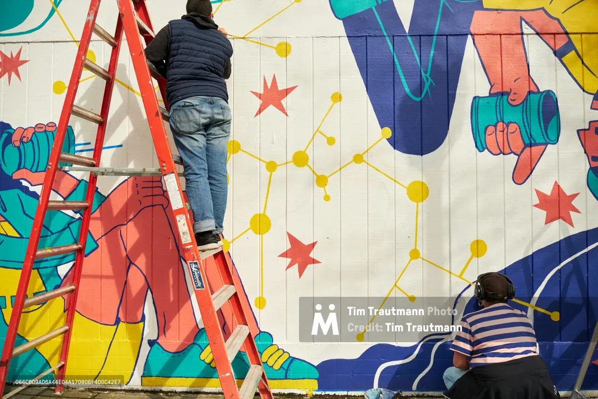 Two artists collaborate on an expansive public mural titled 'Let's Talk' on the exterior wall of Open Signal in Portland's Eliot neighborhood. One artist works from a red ladder, carefully painting details on the white concrete block surface, while another artist in a striped shirt works at ground level with headphones. The vibrant mural features bold geometric hands in coral and blue tones, scattered red stars, and a constellation of golden dots connected by linear pathways across the wall's vertical planks.