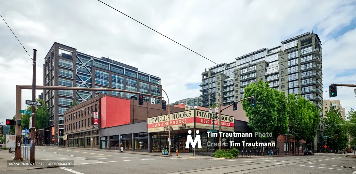 An eerily vacant Burnside Street stretches before Powell's City of Books in Portland's Pearl District during the early days of COVID-19 lockdown. The iconic bookstore's familiar red and cream signage stands sentinel over deserted crosswalks and silent traffic lights, while modern residential towers loom against an overcast Pacific Northwest sky. The absence of pedestrians and vehicles creates a haunting stillness that captures the profound pause that settled over urban America in March 2020.
