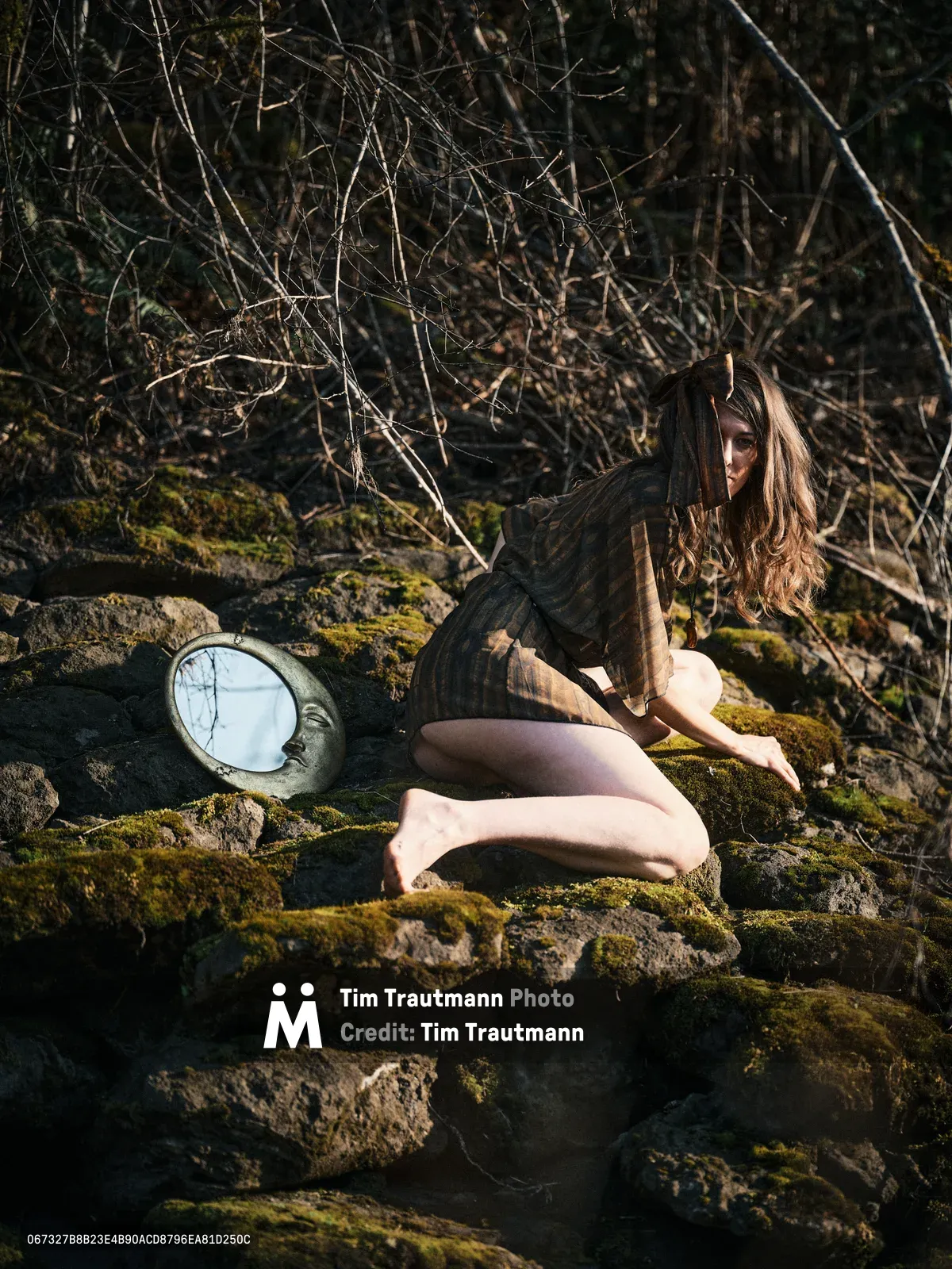 A contemplative nude figure sits gracefully among moss-covered volcanic rocks along Oregon's Floodplain Trail, bathed in dappled forest light. The subject's long auburn hair catches golden rays filtering through bare winter branches, while a circular mirror rests against the verdant stone formations. The intimate scene captures a moment of quiet introspection within the raw beauty of the Columbia River Gorge wilderness.