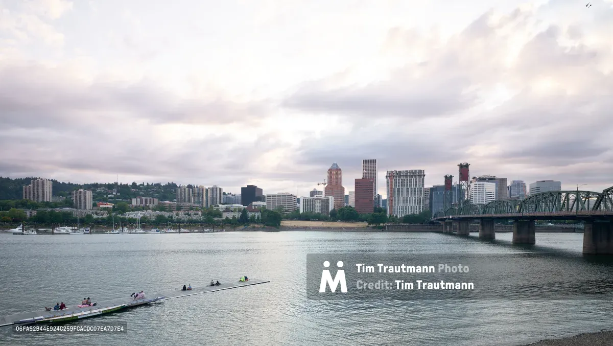 People enjoy leisure time at Holman Dock along the steel-gray waters of the Willamette River, with Portland's downtown skyline rising beyond the water. The city's mix of modern towers and historic buildings is framed by the iconic green steel trusses of the Hawthorne Bridge. The moody afternoon light casts a silvery sheen across the river's surface, while forested hills and residential neighborhoods create a verdant backdrop to the urban core.