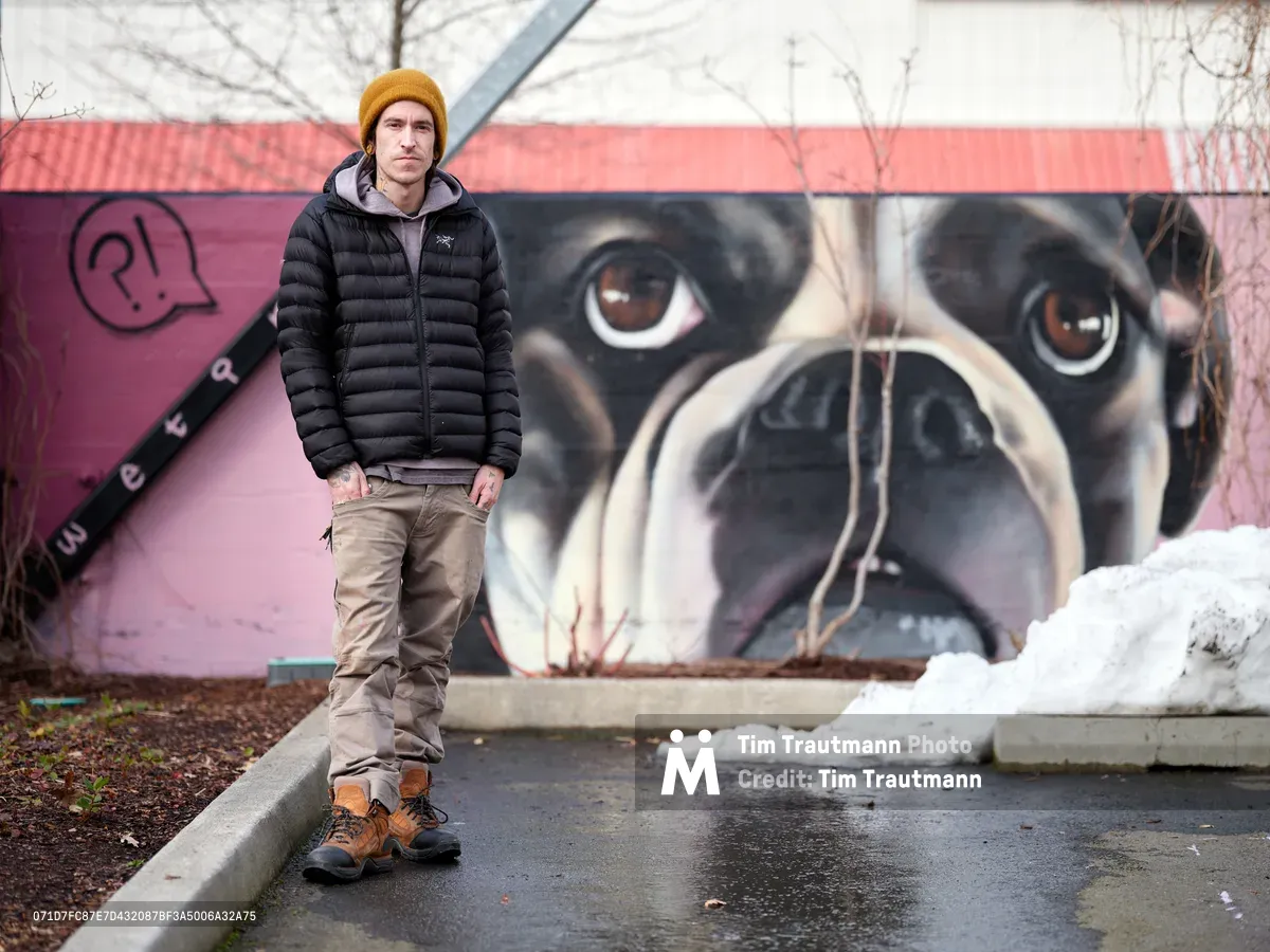 Painter and muralist Wetiko stands confidently before his completed dog portrait mural at the Clay Creative Building in Portland's Hosford-Abernethy neighborhood. The artist, wearing a golden mustard beanie and black puffer jacket, poses against the striking backdrop of his hyperrealistic canine artwork painted on pink-washed concrete walls. Winter's remnants are visible in patches of snow scattered across the wet pavement, while bare tree branches frame the scene under overcast Pacific Northwest skies.