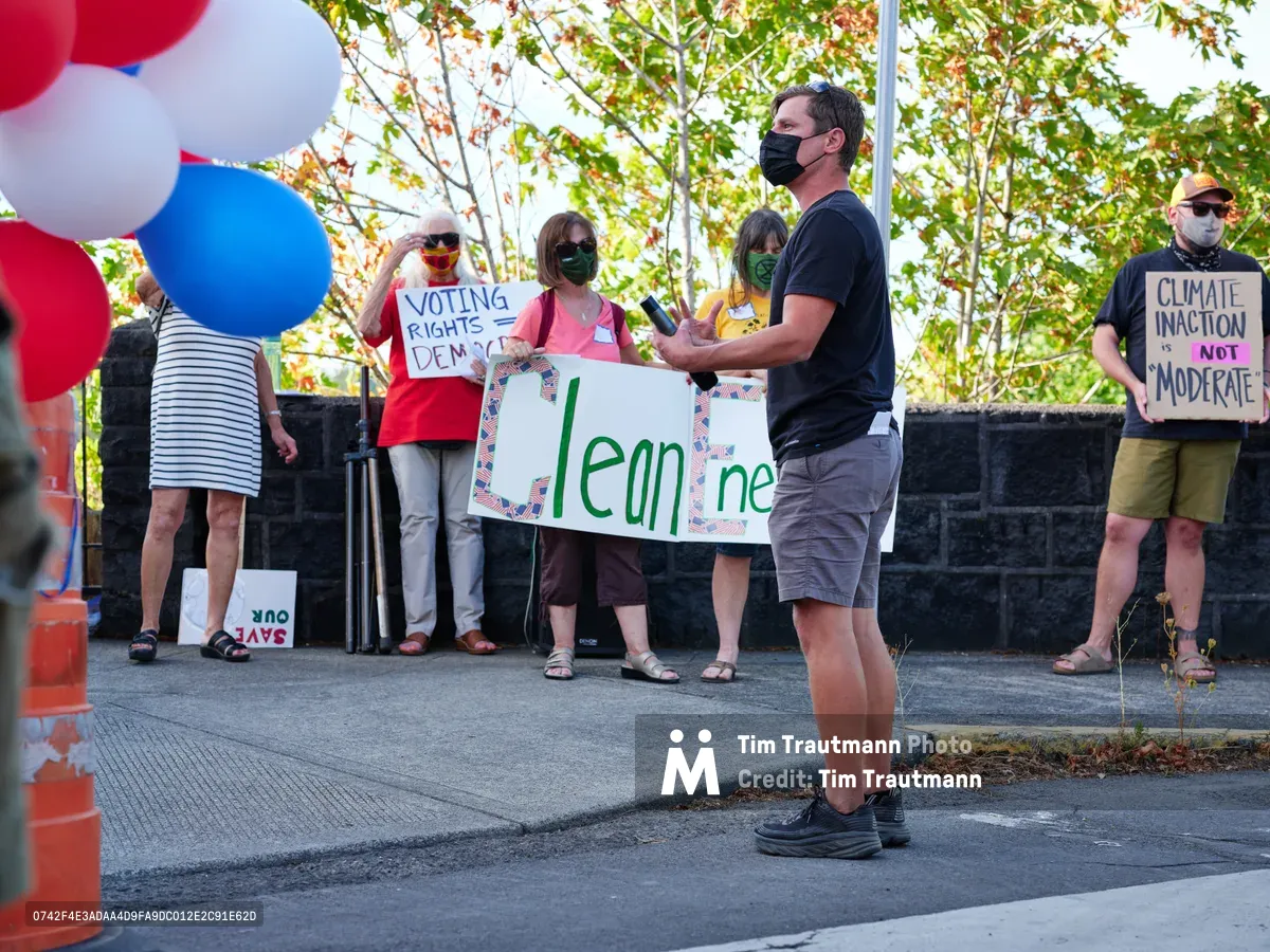 Organizer Michael Gaskill addresses a small gathering of environmental activists outside Representative Kurt Schrader's office on McLoughlin Promenade in Oregon City. Masked protesters hold handmade signs advocating for climate action, including messages about clean energy and democratic voting rights, while patriotic red, white, and blue balloons frame the scene. The afternoon light filters through autumn foliage, casting dappled shadows across the concrete plaza where citizens exercise their right to peaceful assembly.