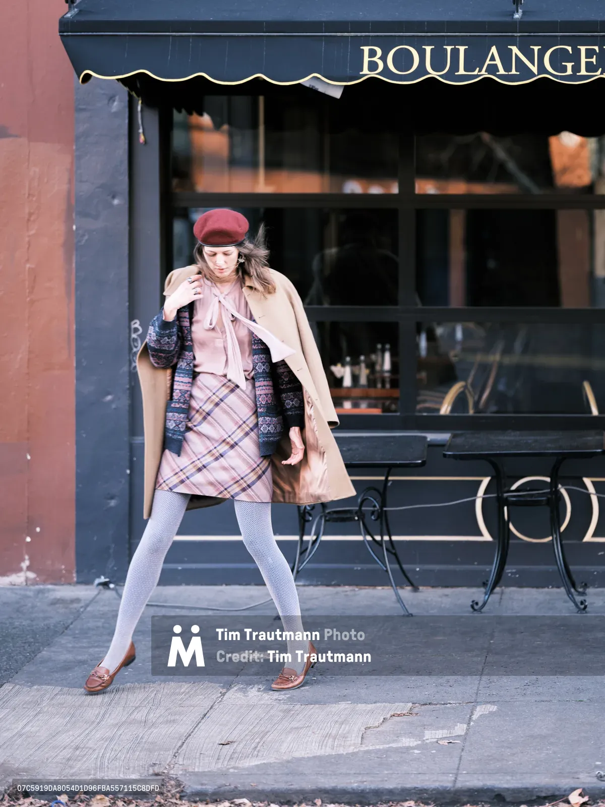 A young woman strides confidently past a French boulangerie on Northeast Alberta Street in Portland, Oregon, embodying effortless street style. She wears a burgundy beret, camel coat draped over her shoulders, plaid mini skirt, and white fishnet stockings, creating a perfect blend of Parisian sophistication and urban Portland charm. The scene captures the golden afternoon light filtering through the storefront windows, with wrought iron café chairs positioned invitingly on the sidewalk. Her purposeful gait and layered autumn ensemble speak to the intersection of European elegance and Pacific Northwest casual culture.