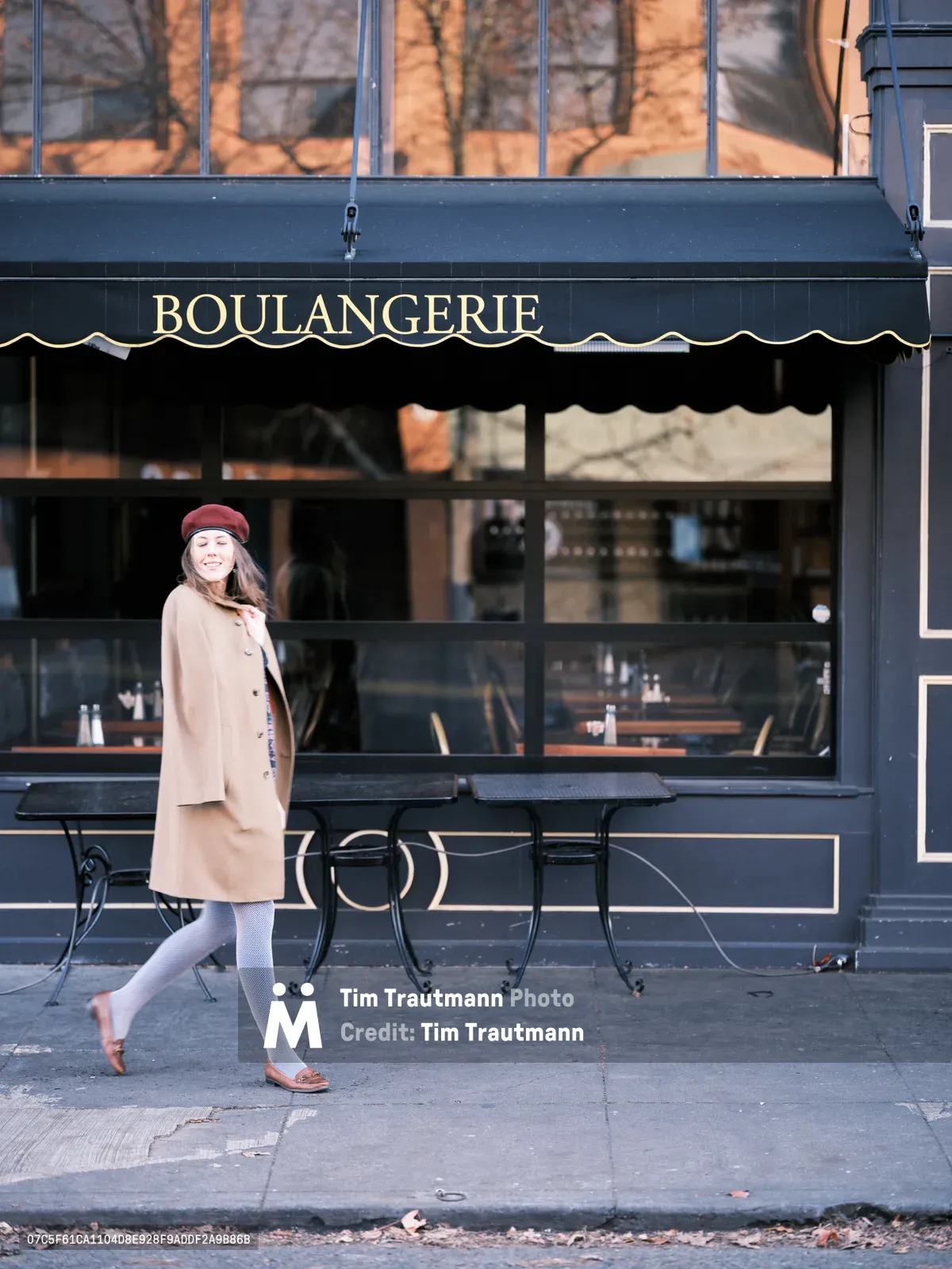 A woman in a camel wool coat and burgundy beret walks gracefully past a charming French boulangerie on Northeast Alberta Street in Portland's Alberta Arts District. Golden morning light bathes the dark navy storefront with its classic awning and large windows, while bare winter branches create delicate shadows above. Her white tights and brown loafers complete the quintessentially Parisian ensemble as she moves through this trendy Portland neighborhood.