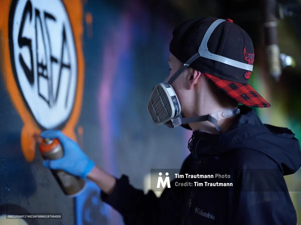 A street artist wearing a Chicago Bulls snapback and protective respirator carefully works on a vibrant mural within the industrial confines of Clay Creative's parking garage in Portland's Hosford-Abernethy neighborhood. The scene captures the intersection of urban art and workspace, as colored lighting bathes the concrete walls while the artist methodically applies paint to their canvas. The atmospheric garage setting transforms into a creative sanctuary, where spray paint cans and artistic vision merge under artificial illumination.