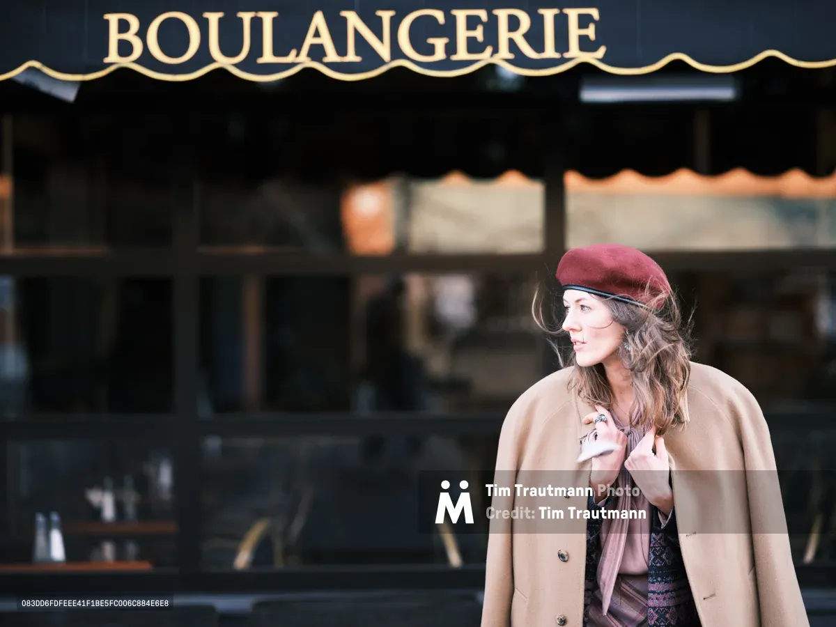 A woman in a burgundy beret and camel-colored coat stands contemplatively outside a traditional French boulangerie on Alberta Street in Portland, Oregon. The golden afternoon light catches her profile as she gazes thoughtfully to the side, her wavy brown hair framing her face beneath the classic French hat. The dark green storefront with ornate gold lettering creates an authentic European bakery atmosphere, while the shallow depth of field draws focus to her elegant silhouette against the blurred urban backdrop.