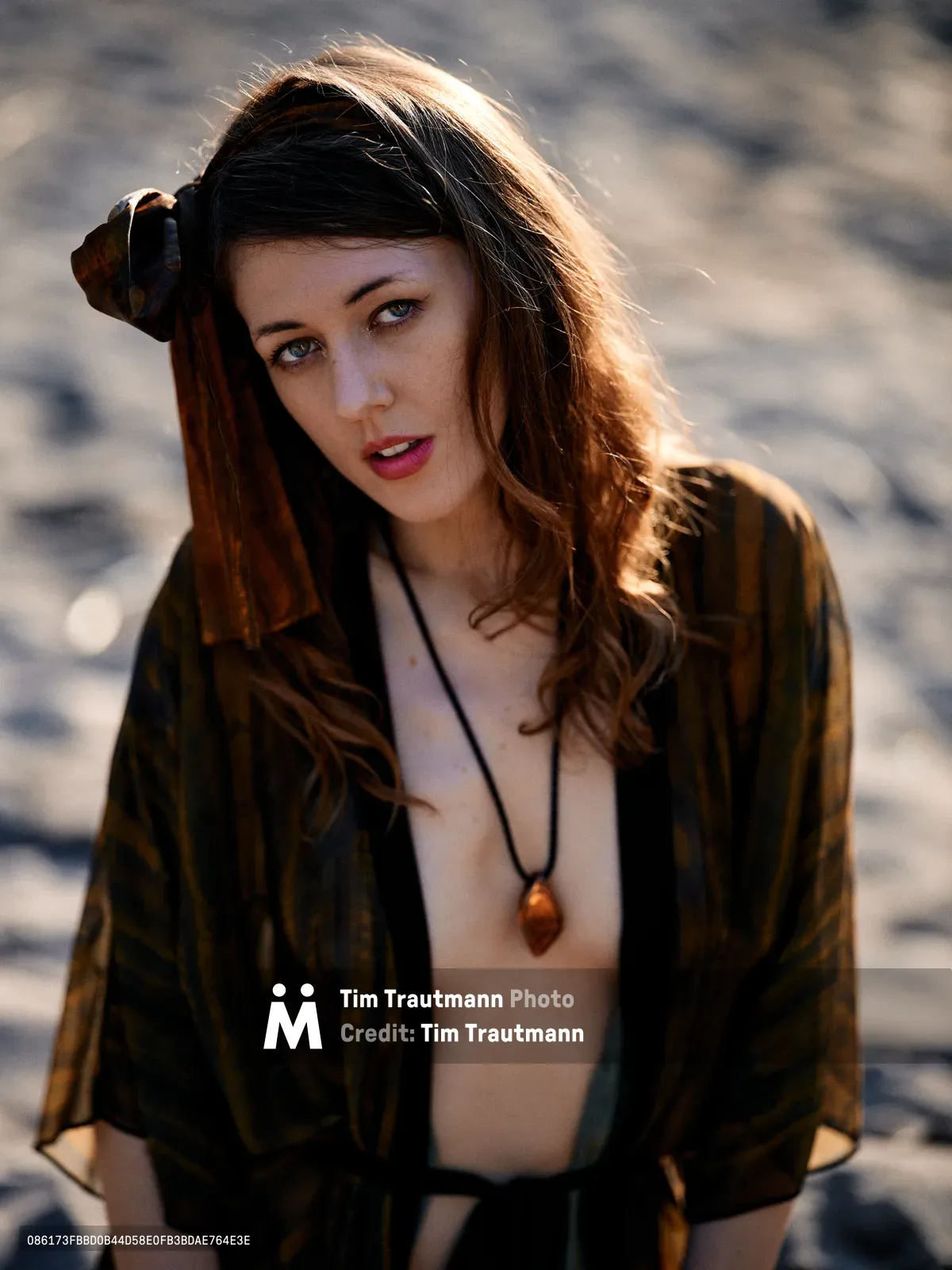 A young woman with flowing brown hair catches warm golden sunlight along the Floodplain Trail in Multnomah County, Oregon. She wears an olive military-style jacket over a simple top with an amber pendant necklace, her hand gracefully positioned near her hair as she gazes directly at the camera. The soft evening light creates a luminous halo effect around her silhouette against the blurred natural backdrop of the river valley landscape.