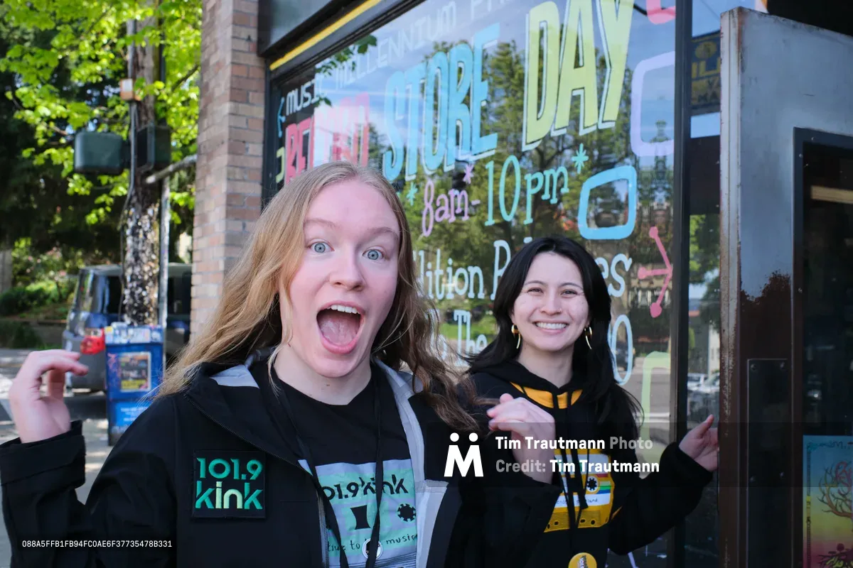 Two young women radiate pure joy and excitement outside Music Millennium record store on East Burnside Street in Portland, Oregon. The blonde woman in front displays an exaggerated expression of surprise and delight, her mouth wide open and eyes bright, while her brunette companion smiles warmly behind her, both wearing branded radio station apparel. The scene captures a moment of spontaneous enthusiasm against the backdrop of the iconic independent record store's large storefront windows, with lush green spring foliage framing the urban setting.