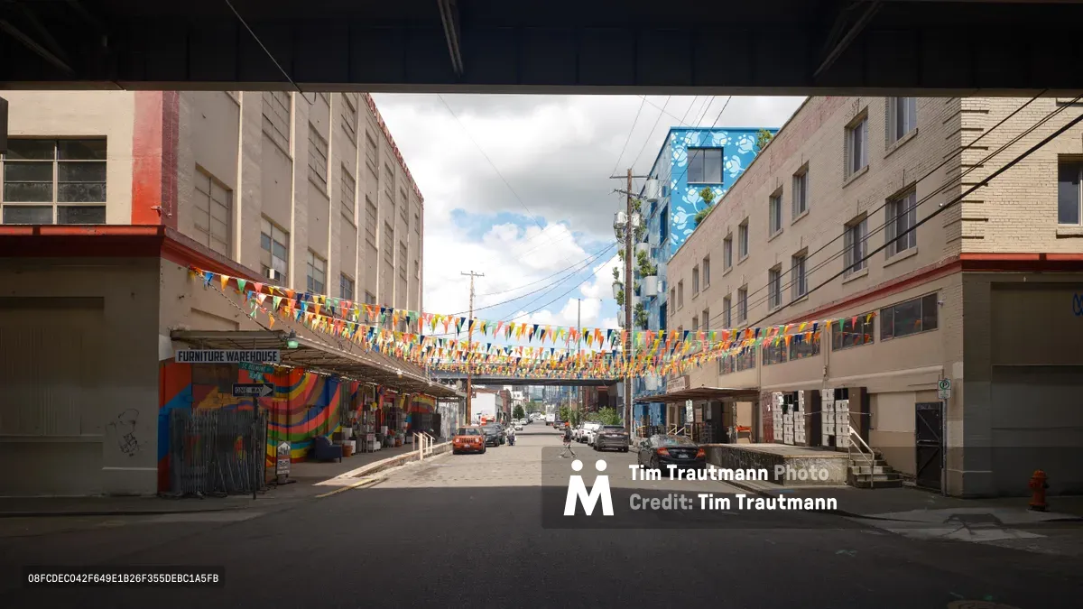 Colorful triangular bunting stretches across a working-class Portland street, creating a celebratory canopy between mid-century apartment buildings and warehouses. The vibrant pennant flags in red, blue, yellow, and green contrast against the utilitarian concrete and brick architecture of the City Liquidators district. Afternoon light filters through scattered clouds, illuminating the festive decorations that temporarily transform this industrial corridor into a community celebration space.