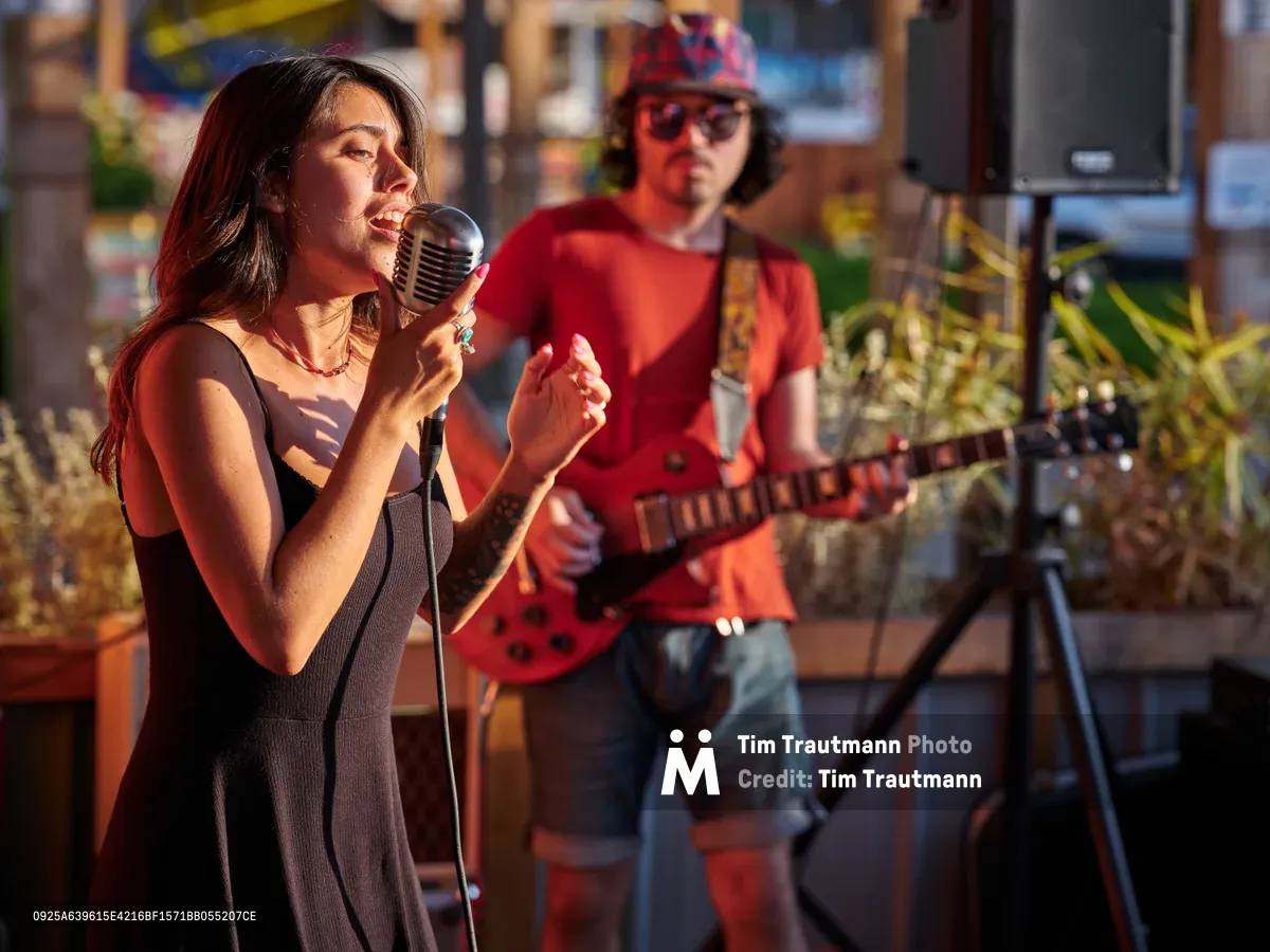 Vocalist Aubrey Gordon commands the vintage microphone with passionate intensity as golden late-afternoon light bathes her performance at Portland Mercado in Southeast Portland. Behind her, a guitarist in red flannel and patterned cap provides accompaniment, while tropical plants and the market's vibrant atmosphere create an intimate outdoor concert setting. The warm, amber lighting transforms this community gathering space into a stage where music and neighborhood culture converge.