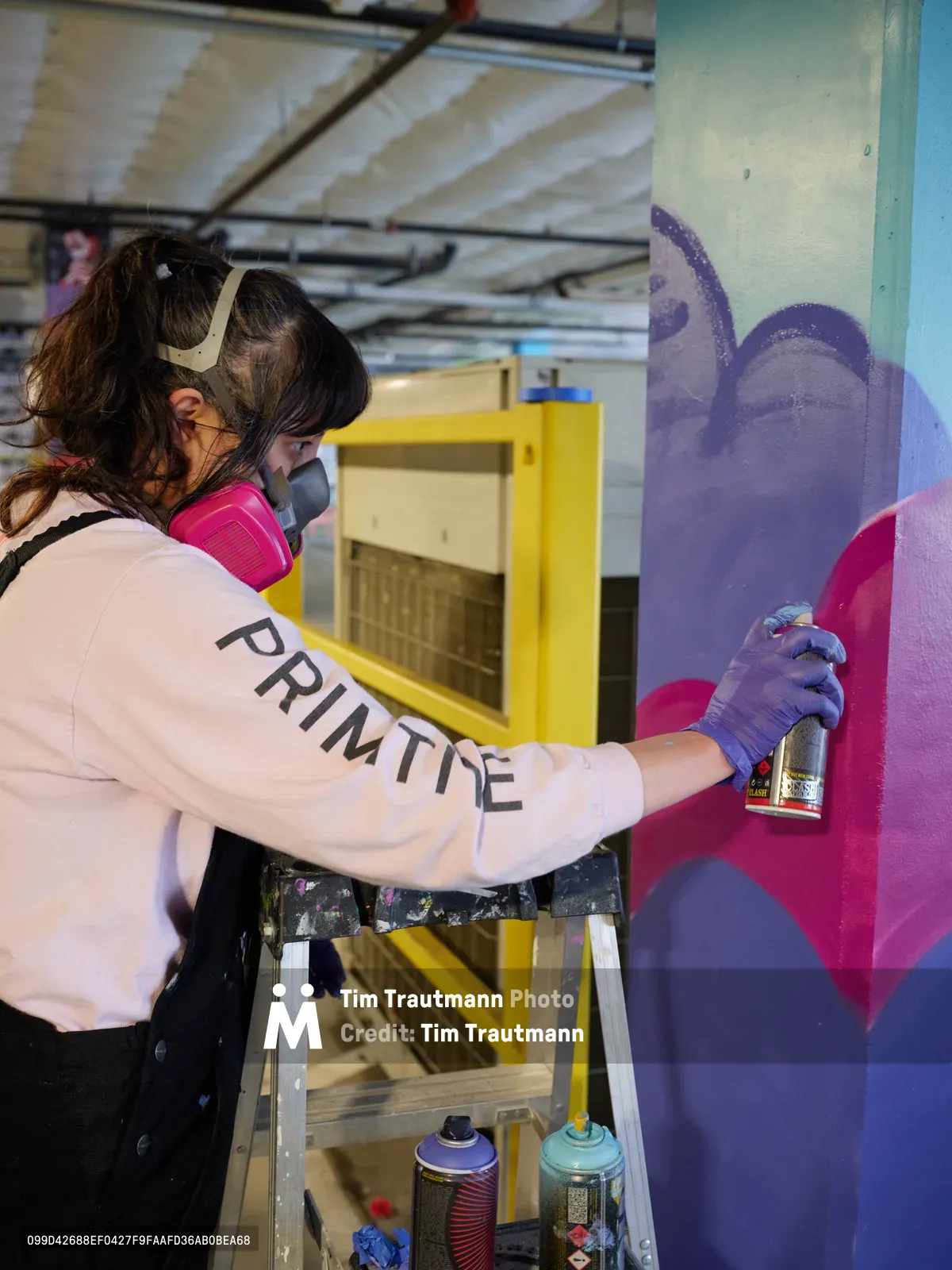 A focused artist wearing protective gear and a white 'PRIMTE' shirt applies vibrant purple spray paint to an emerging mural on a concrete wall. The industrial setting of Clay Creative's parking garage in Southeast Portland provides raw backdrop of exposed ceiling beams and yellow storage units. Her concentrated posture and precise movements capture the intimate process of transforming urban space into art, while spray cans rest on a wooden stepladder beside her.