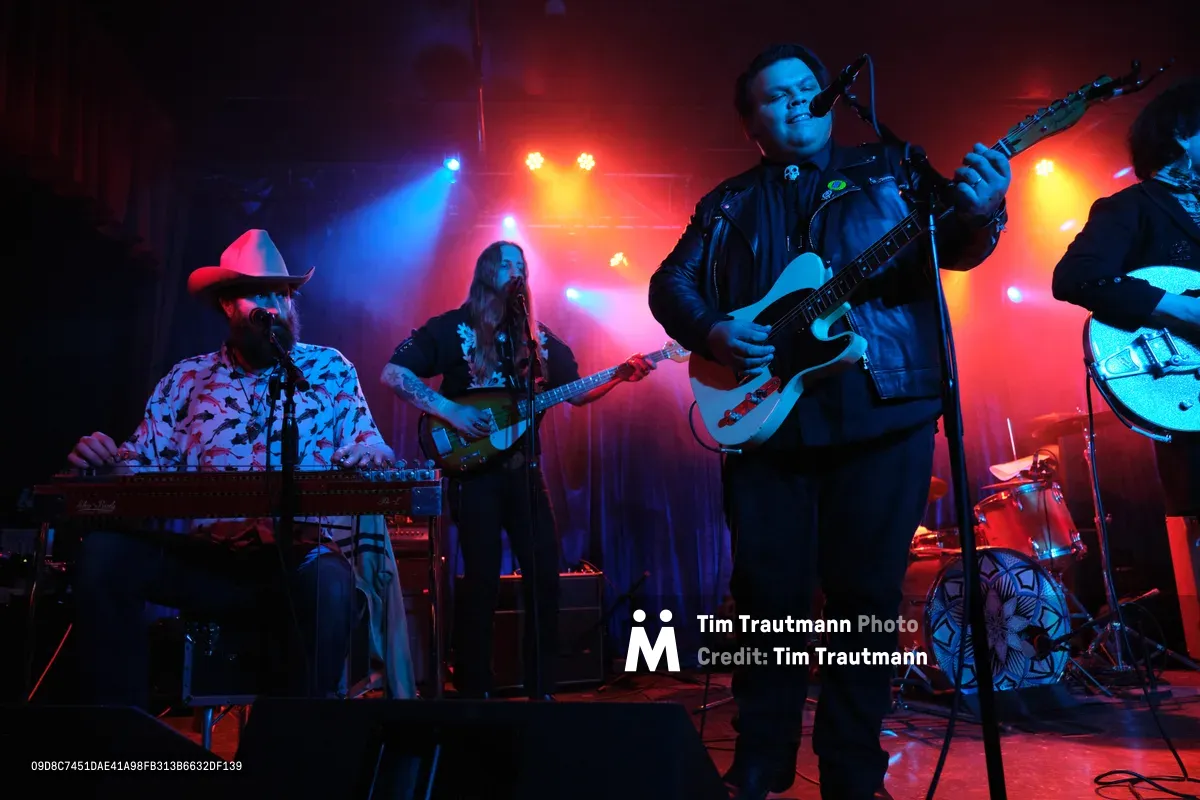 Barry Walker, Tobias Berblinger, and Victor Franco of Roselit Bone command the intimate stage at Dante's in Portland, their silhouettes carved against a wash of crimson and azure stage lights. Walker's cowboy hat catches the glow as he works the synthesizer, while Franco's leather jacket gleams under the dramatic lighting as he grips his white electric guitar. The atmospheric haze and moody lighting create an electric tension that defines Portland's underground music scene.