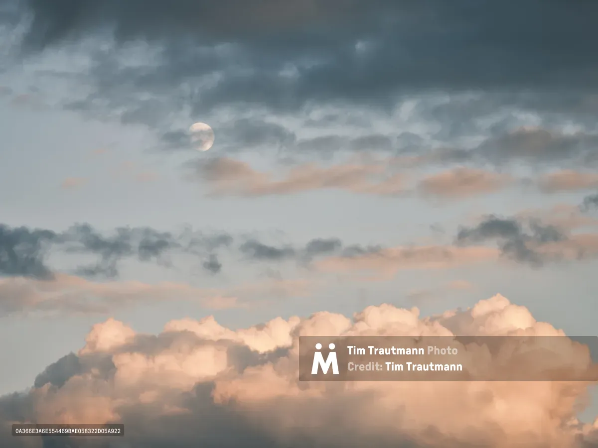 A pale moon emerges through layers of dramatic storm clouds above the Clark County Fairgrounds in Vancouver, Washington. The twilight sky creates a theatrical backdrop of steel-gray and rose-tinted cumulus formations, with the moon serving as a luminous focal point amid the turbulent atmosphere. The layered cloudscape suggests the approaching dusk of a fair evening, where nature's own spectacle competes with the festivities below.