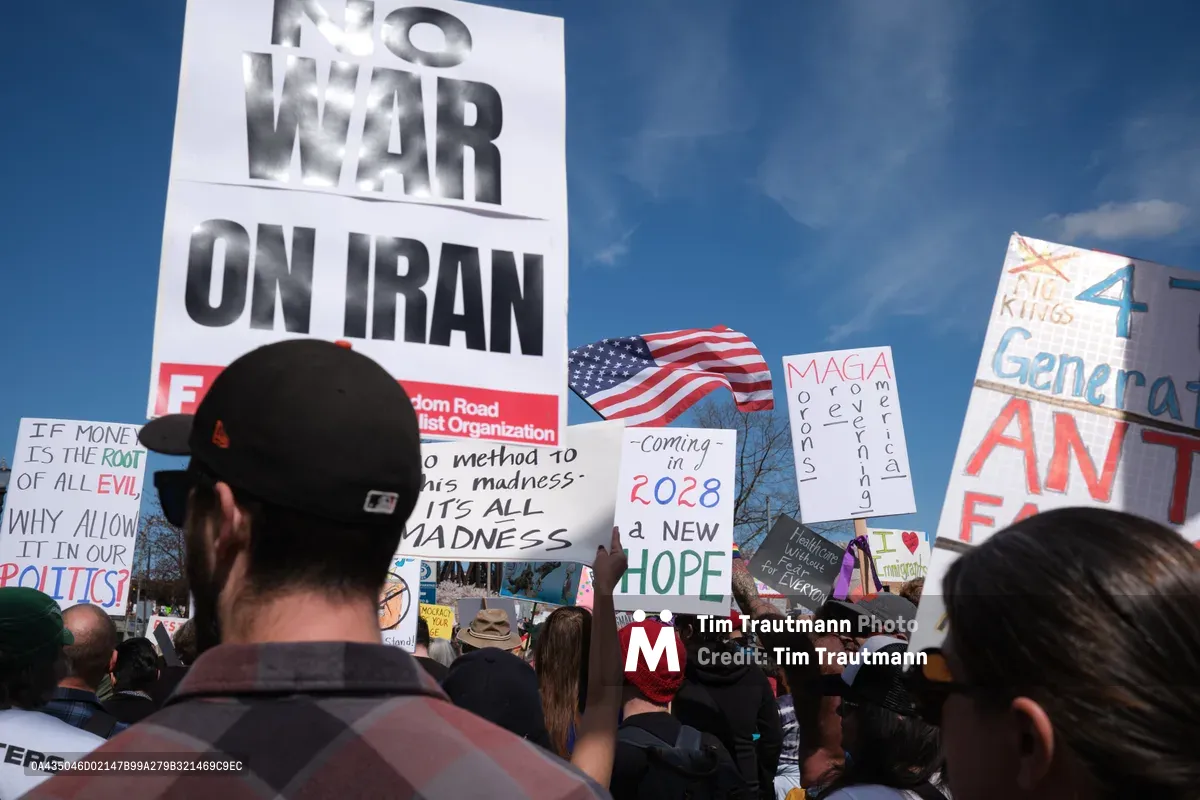 Protesters gather beneath a crystalline blue sky in Portland's historic Old Town district, their handmade signs creating a mosaic of dissent against potential military action. The crowd stretches into the distance along Southwest Ankeny Street, with prominent placards declaring "NO WAR ON IRAN" and references to 2028 hope rising above a sea of baseball caps and winter jackets. An American flag waves paradoxically among the anti-war messages, while demonstrators of various ages stand shoulder-to-shoulder in peaceful assembly.