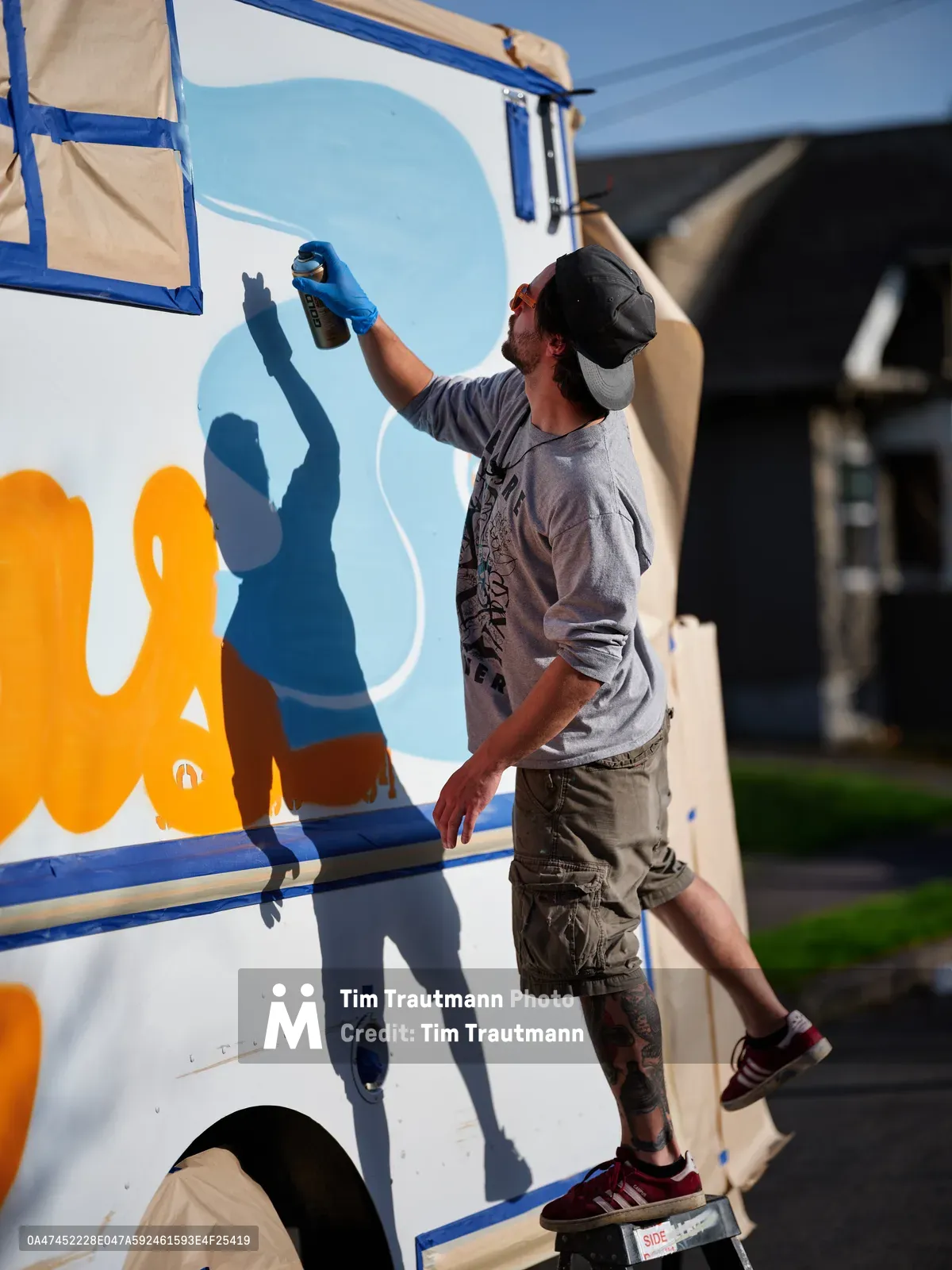 A tattooed muralist in a baseball cap and gray t-shirt reaches upward with a blue spray can, adding vibrant orange and blue elements to a white food truck's exterior. Sharp afternoon shadows dance across the vehicle's surface as the artist works methodically on the evolving mural. The urban scene unfolds on Portland's Northeast 9th Avenue, where residential architecture forms a soft backdrop to this creative transformation.