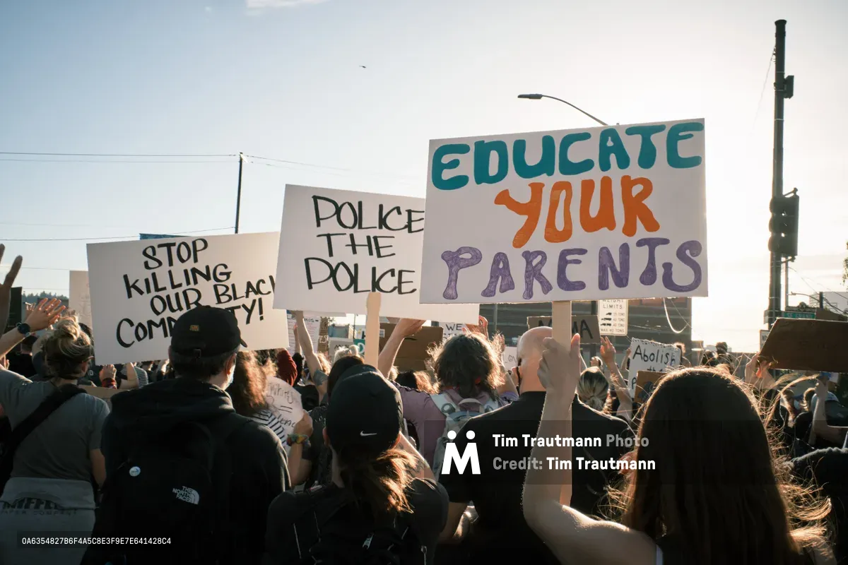 A dense crowd of protesters moves purposefully across Portland's Morrison Bridge, their handmade signs catching the golden hour light against a pale sky. The demonstrators hold messages including "STOP KILLING OUR BLACK COMMUNITY," "POLICE THE POLICE," and "EDUCATE YOUR PARENTS," their colorful lettering standing out against white poster board. The late afternoon sun backlights hair and shoulders, creating an atmosphere of determined solidarity as the group advances toward downtown Portland. Power lines and urban infrastructure frame the scene, emphasizing the grassroots nature of this civil rights demonstration.