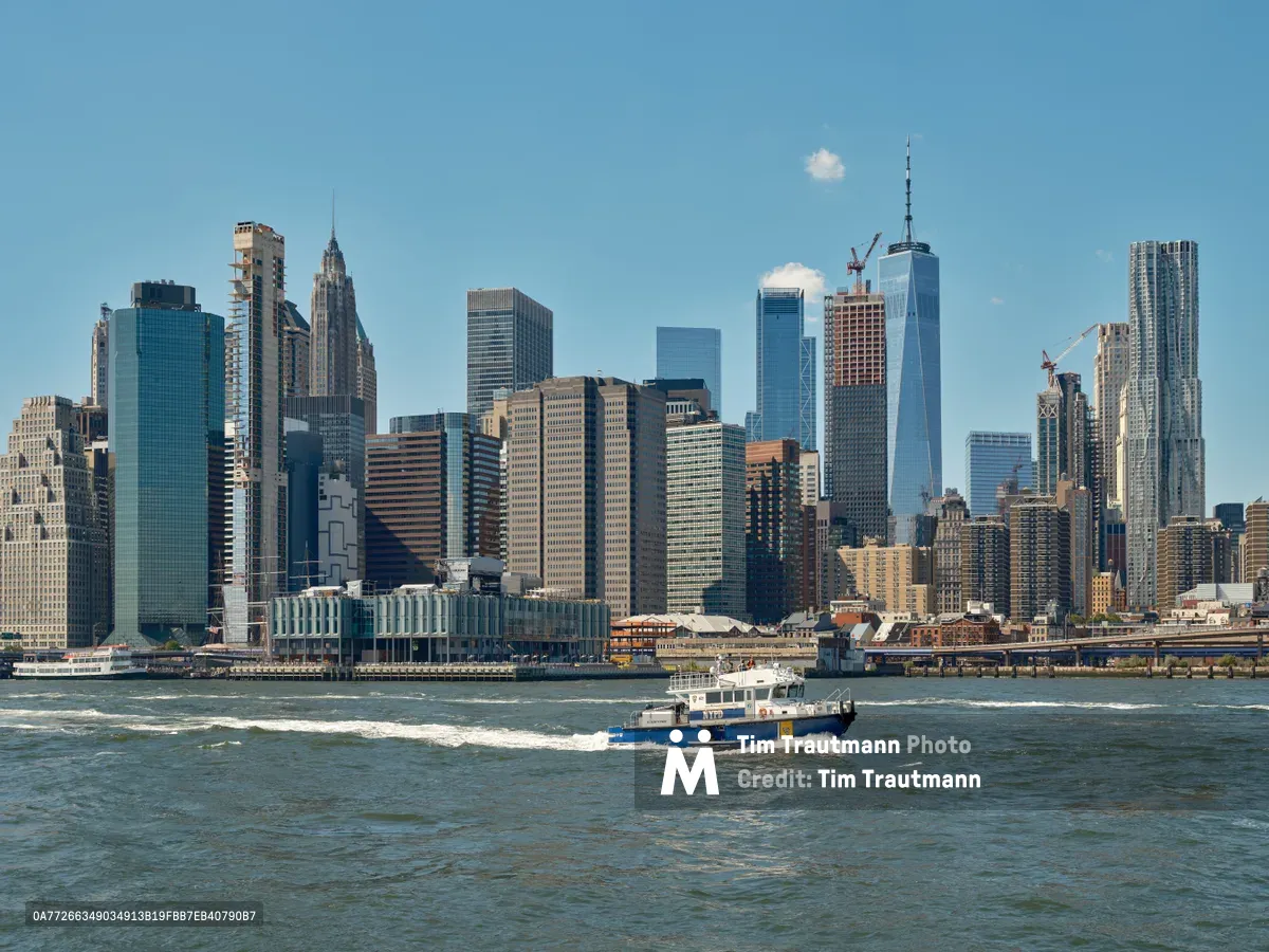 An NYPD patrol boat cuts through the choppy waters of New York Harbor, creating white wake trails as it patrols near the towering Financial District skyline. The crystalline afternoon light illuminates the iconic Manhattan silhouette, from the Art Deco spire of the Woolworth Building to the gleaming glass facades of One World Trade Center. The maritime scene captures the daily rhythm of harbor security operations against the dramatic backdrop of America's premier financial center, photographed from the historic Fulton Ferry Landing waterfront.