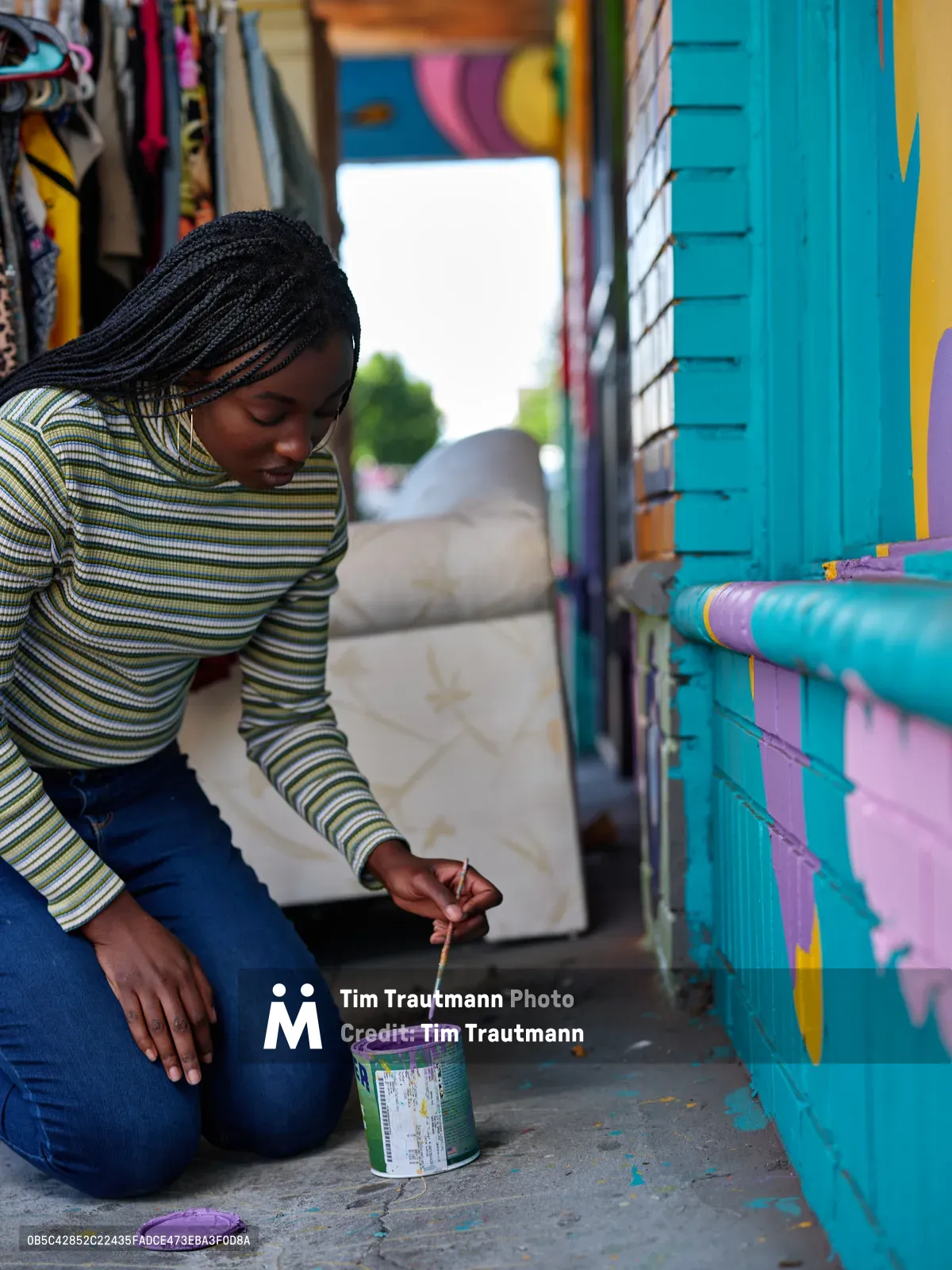 Naomi Likayi kneels on the sidewalk, carefully signing her vibrant turquoise and pink mural with purple paint in Portland's Alberta district. The intimate moment captures the artist's focused concentration as she puts the finishing touches on her colorful street art, surrounded by the creative energy of this Northeast Portland neighborhood. Natural daylight illuminates her striped sweater and braided hair as she works with steady precision, paint can beside her on the concrete.