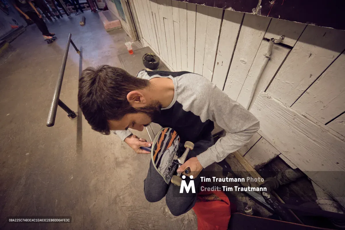 A young craftsman hunches over a skateboard deck in the industrial confines of Commonwealth Skateboarding on Southeast 20th Avenue in Portland's Buckman neighborhood. Warm overhead lighting casts dramatic shadows across weathered concrete floors as he meticulously adjusts the board's components, surrounded by the authentic clutter of skate tools and equipment. The intimate scene captures the meditative ritual of skateboard maintenance, where precision meets passion in this legendary Portland skate sanctuary.