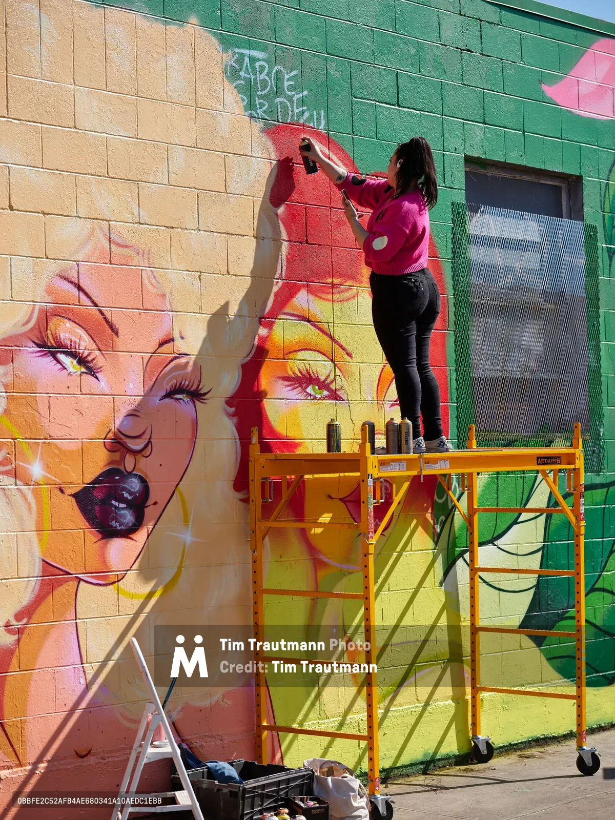 Multidisciplinary artist Vilatose works from a yellow scaffolding platform, spray painting vivid details on a large-scale portrait mural adorning a brick wall in Portland's Alberta Arts District. The emerging artwork features luminous skin tones, piercing green eyes, and bold red elements against the textured masonry canvas. Golden afternoon light casts dramatic shadows across the colorful work-in-progress, while paint supplies and equipment scatter below on the concrete sidewalk.