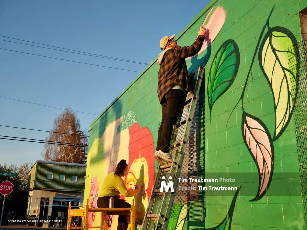 Multidisciplinary artist Vilatose works from a ladder to paint flowing elements on the vibrant green mural 'Jardín Cubana' along Northeast Alberta Street in Portland's historic arts district. Golden hour light bathes the scene as a second artist collaborates below, their coordinated efforts bringing life to the emerald wall depicting three female figures with stylized elements and coral-pink accents. Power lines cross the clear blue sky above this creative transformation in the heart of Portland's cultural corridor.