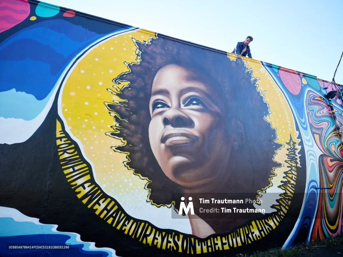 Travis Fields stands triumphantly atop the building beside his completed mural of poet Amanda Gorman in Portland's Alberta Arts District. The large-scale portrait depicts Gorman with a luminous golden halo framing her natural hair, her face rendered in warm earth tones with an upward gaze of hope and determination. The surrounding wall bursts with psychedelic swirls of magenta, cyan, and electric blue, creating a cosmic backdrop that celebrates both the subject and the vibrant street art culture of Northeast Portland.